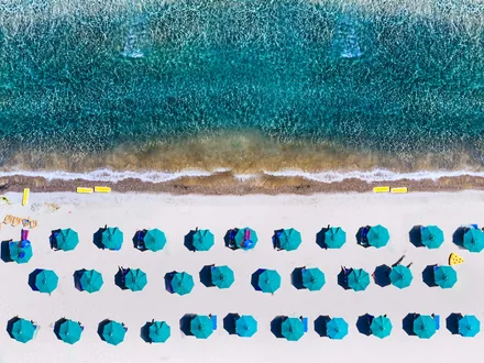 Rader med blå parasoller på stranden sett ovenfra mot turkist vann