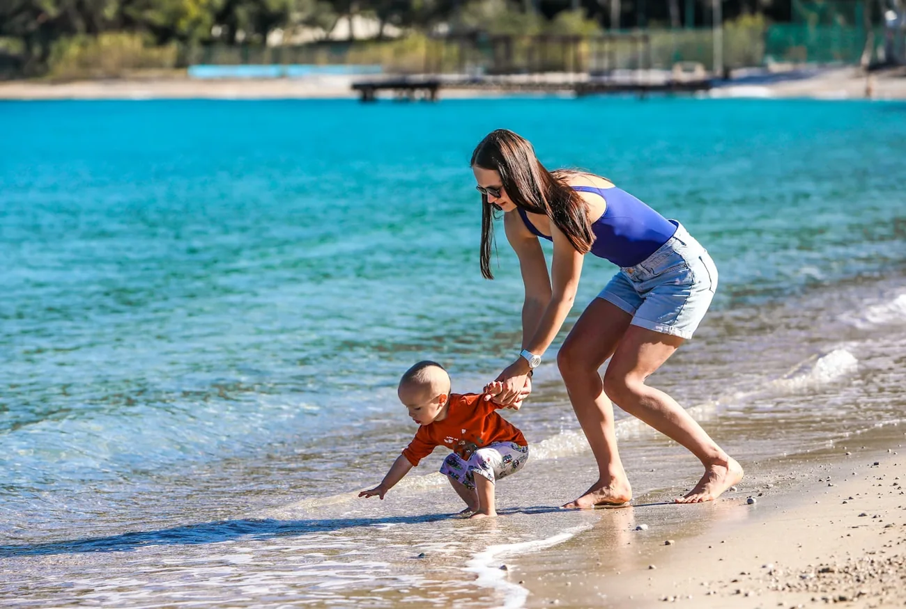 En mor hjelper et lite barn i vannkanten på en tyrkisk strand med turkist vann